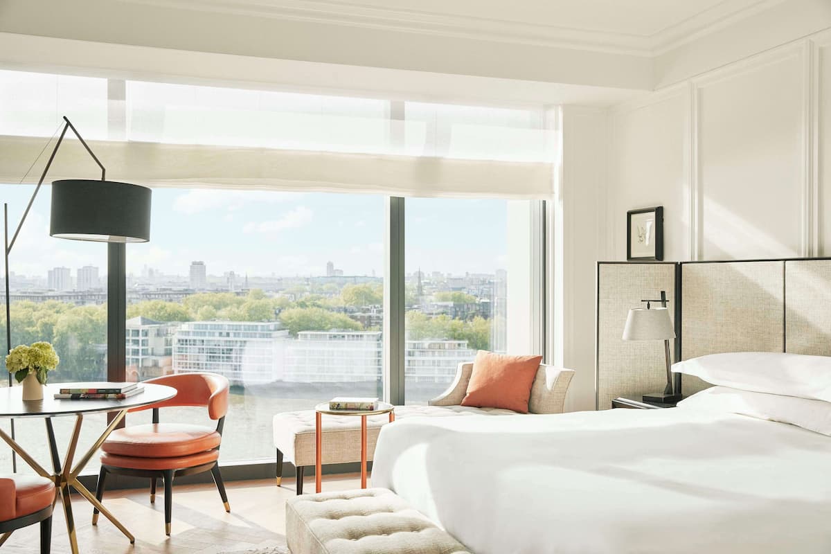 Ein modernes Hotelzimmer mit großem Fenster und Blick auf die Stadt, einem Bett mit weißer Bettwäsche, einem Schreibtisch mit Lampe und einem Sessel.