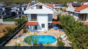 Ein modernes, zweistöckiges Gebäude mit Pool auf dem Dach, umgeben von Wohnhäusern und einer hügeligen Landschaft.