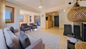 Ein geräumiges Wohnzimmer mit einer Couch, Sesseln, einem gemusterten Teppich und einer Küchenzeile mit Herd und Schränken.