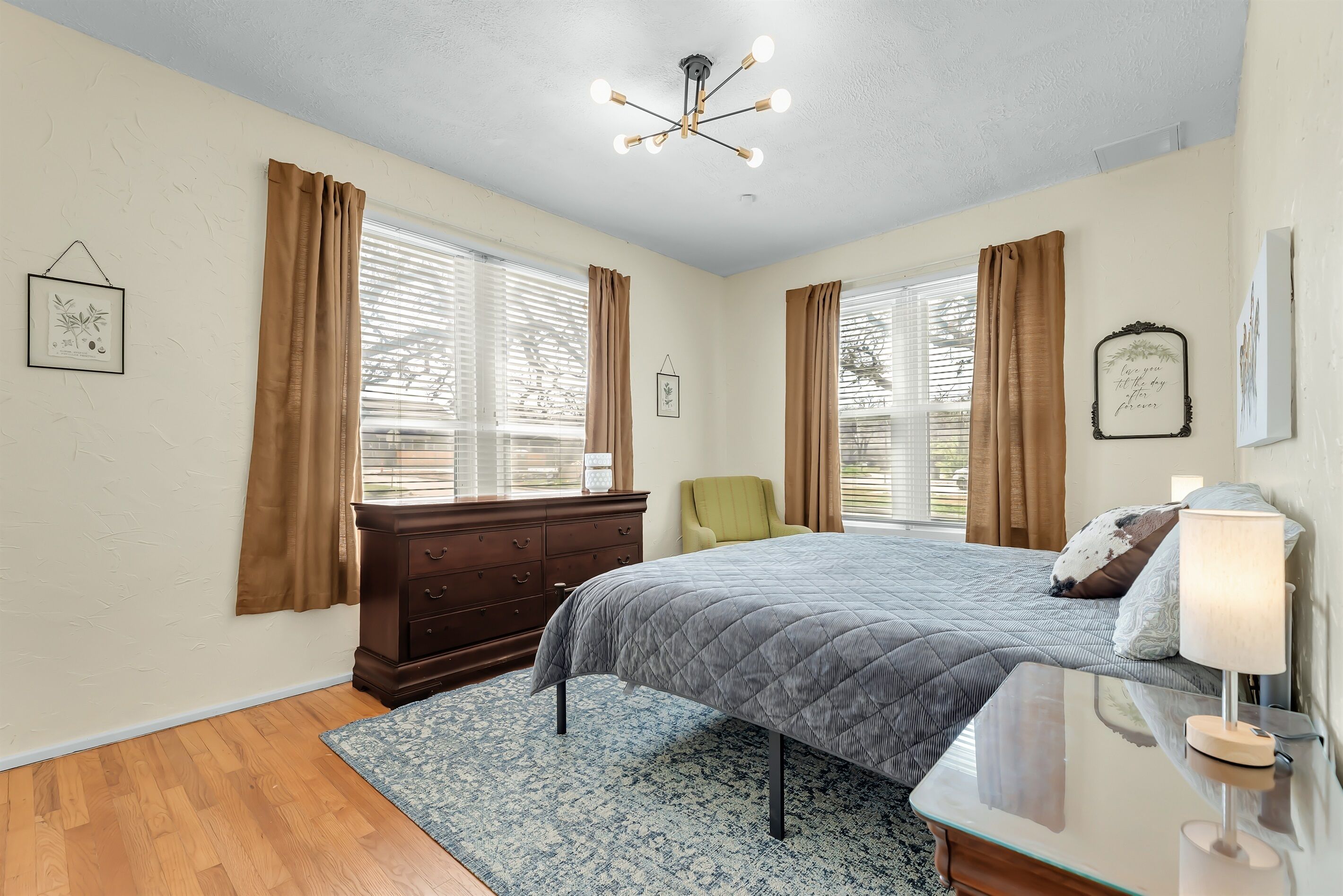 A bedroom with a bed, a dresser, a chair, a desk with a lamp, and a window with curtains.
