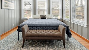 Un dormitorio con una cama grande, un banco y tres ventanas con persianas.