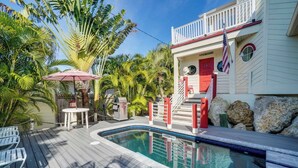 A pool area with a wooden deck, lounge chairs, a small table with an umbrella, and a building with a red door and American flag. - Americana AMI Lighthouse 3BR w/pool + rooftop deck (Holmes Beach)