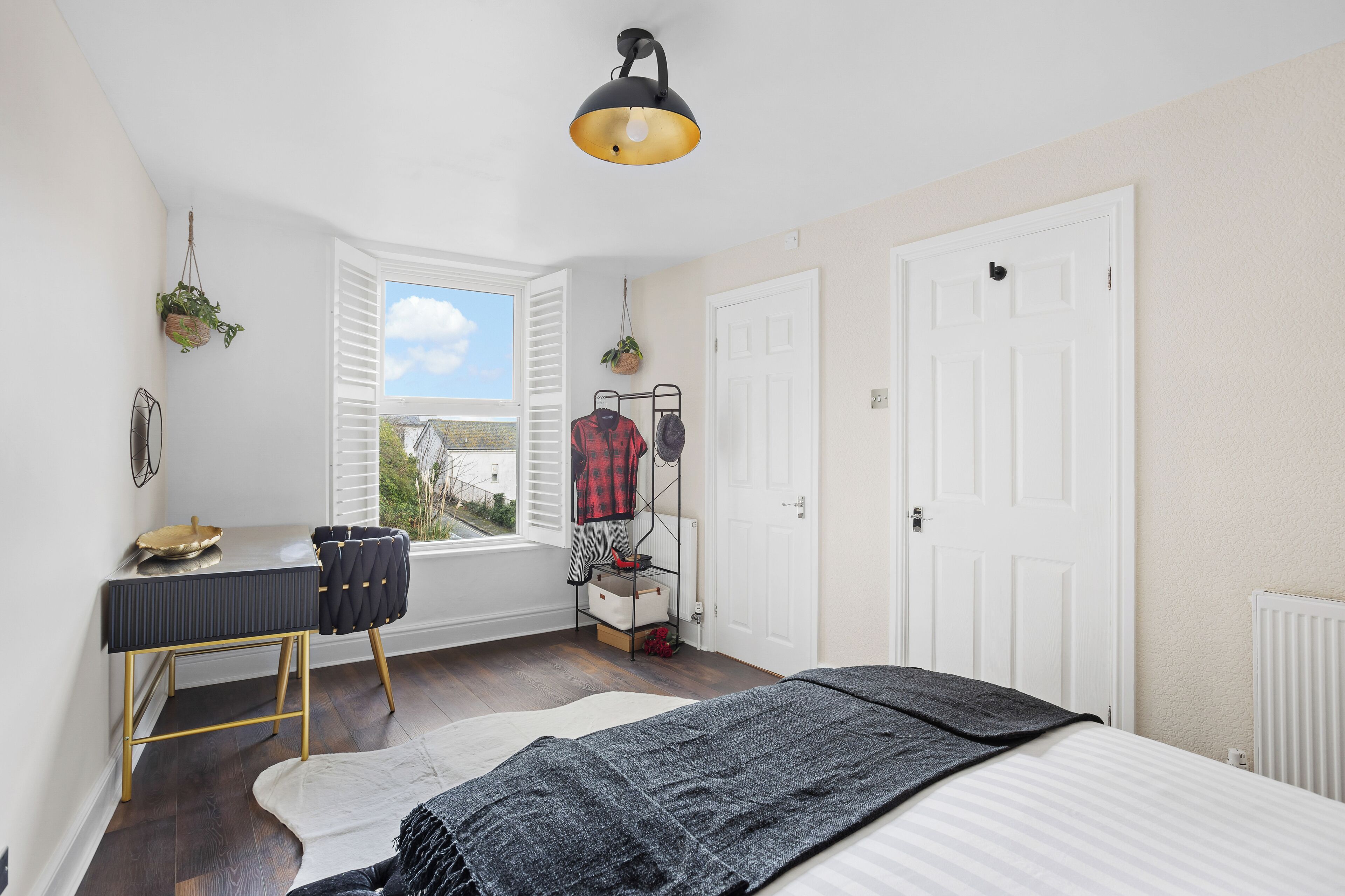 Ein Schlafzimmer mit einem Bett, einem Schreibtisch mit Stuhl, einer hängenden Pflanze und einem Fenster mit Jalousien.