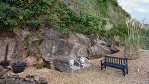 Un banc et deux chaises sont placés sur une surface de gravier, près d’une falaise rocheuse couverte de végétation.