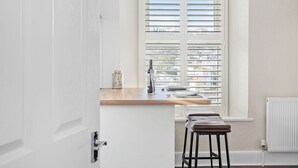 Une cuisine avec un plan de travail en bois, un évier et une fenêtre avec des stores. On y trouve deux tabourets et un radiateur.