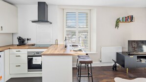 Une cuisine moderne avec des meubles blancs, un plan de travail en bois, une cuisinière encastrée et une grande fenêtre équipée de stores.