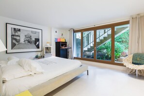 Relax in a serene space with a large bed, framed art, and a desk with a lamp, offering a peaceful view of the outdoor area with lush greenery.