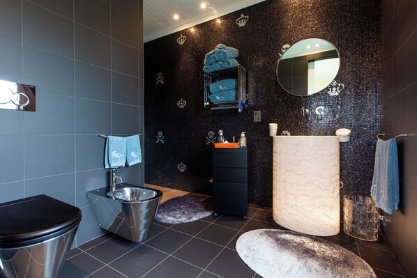 A modern bathroom with a black tiled wall, a large mirror, a sink with a marble countertop, and a toilet with a metallic finish. - Magic Forte (Forte dei Marmi)