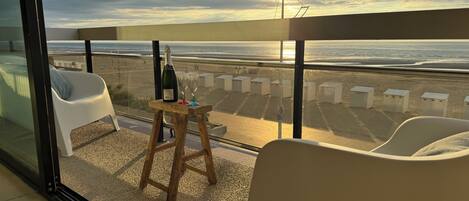 A balcony with a view of the beach, a bottle of champagne, and two glasses on a small stool.