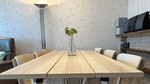 Un coin repas avec une table en bois, six chaises, un vase avec des plantes, une lampe et un mur en briques agrémenté d’une étagère.
