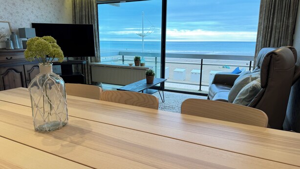 Une chambre d’hôtel moderne avec une table à manger en bois, un téléviseur et une grande fenêtre donnant sur la plage et l’océan.