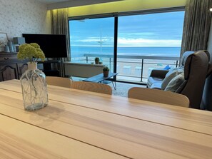 A modern hotel room with a wooden dining table, a TV, and a large window offering a view of the beach and ocean. - Suite Baptiste. Beachfront (Koksijde)