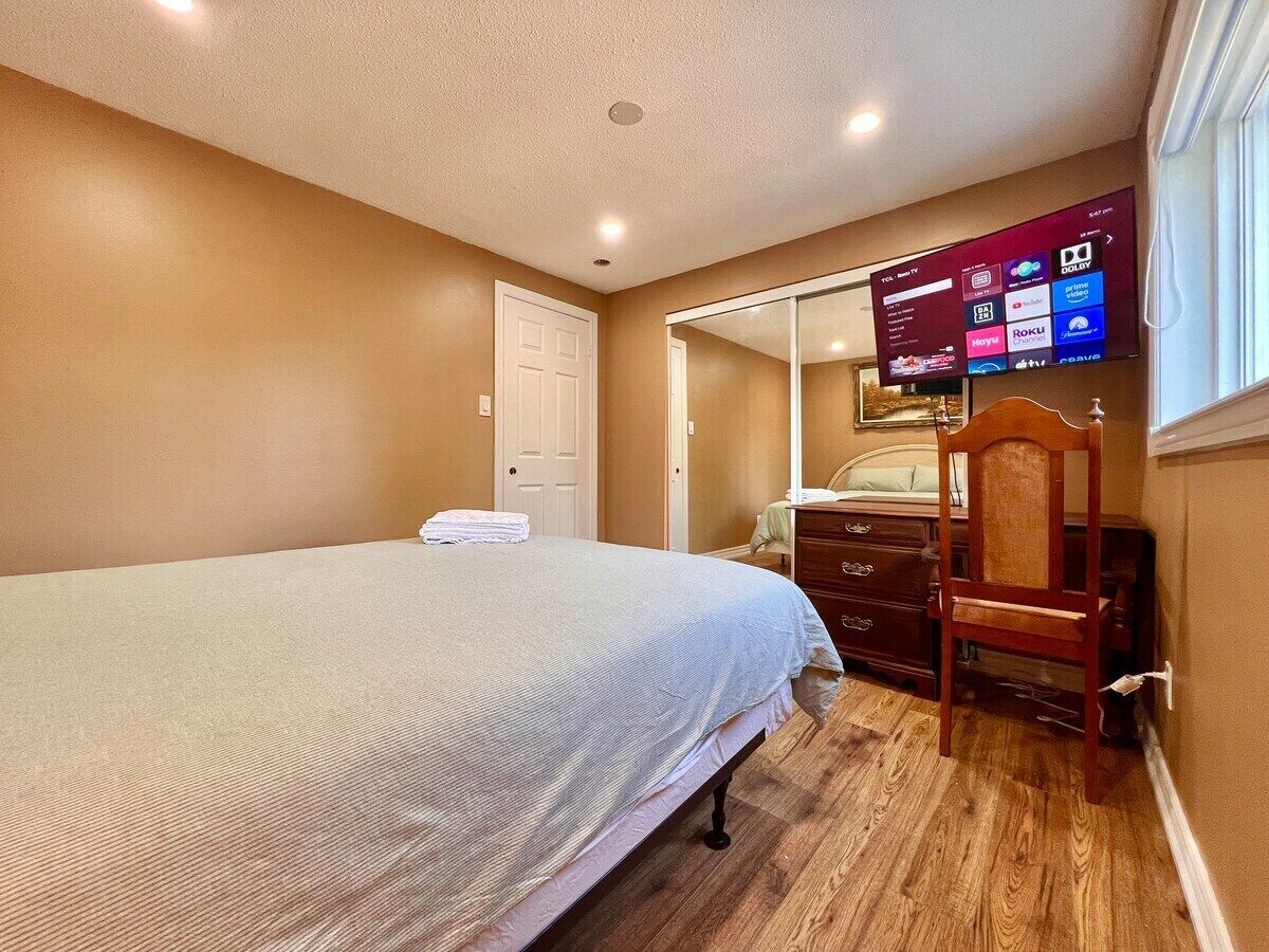 A bedroom with a bed, a wooden dresser, a chair, and a TV mounted on the wall displaying various channels.