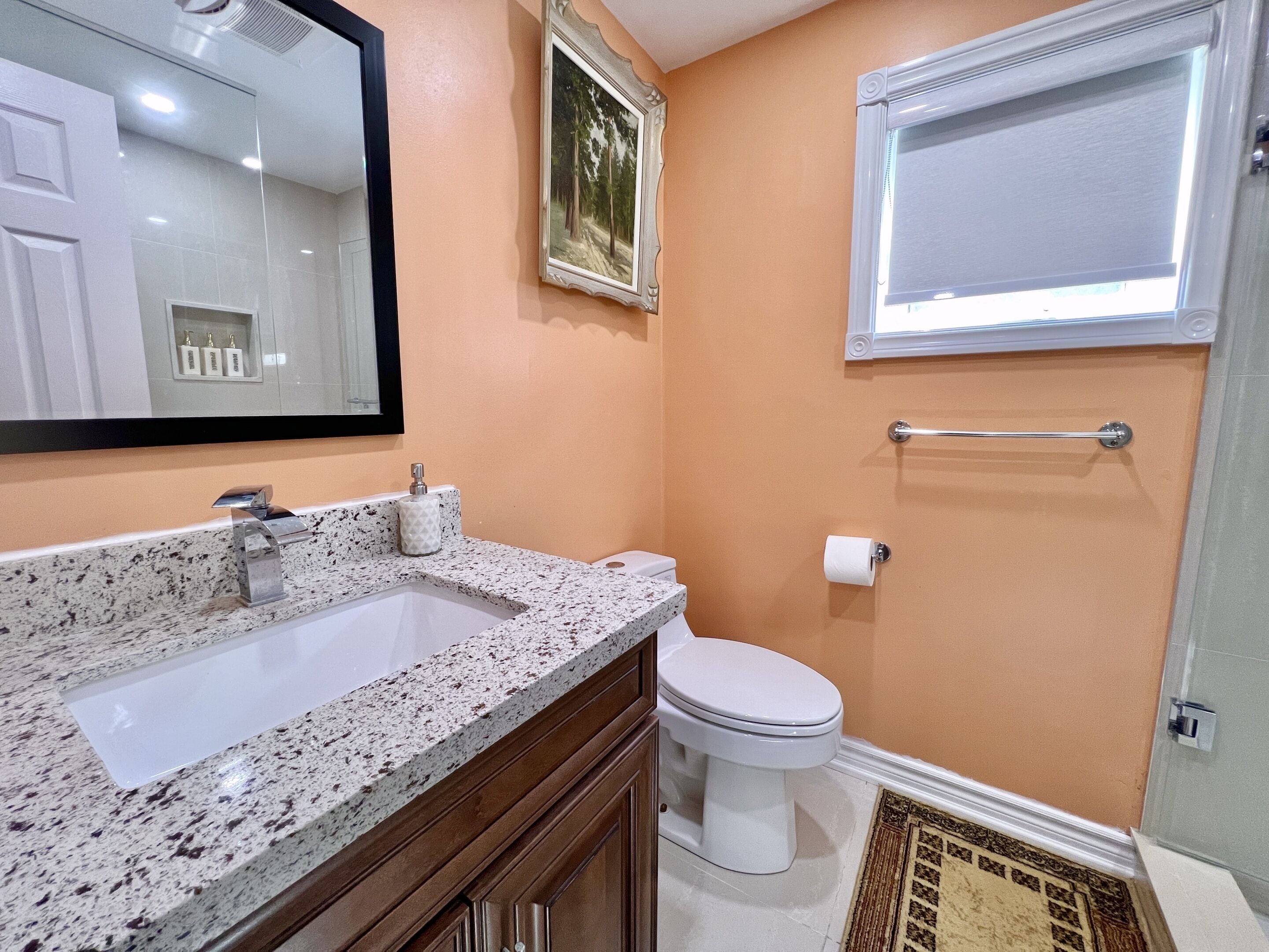 A bathroom with a granite countertop sink, a white toilet, a framed picture on the wall, and a window with a blind.
