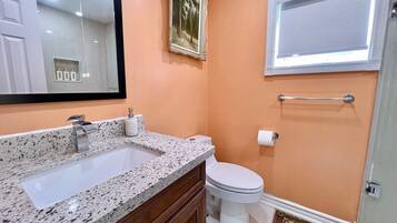 A bathroom with a granite countertop sink, a white toilet, a framed picture on the wall, and a window with a towel bar.