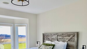A bedroom with a large bed, a wooden headboard, a nightstand with a lamp, and a view of a green field through the window. - The Bavaria Farmhouse - 30 Acre Estate (Chaska)