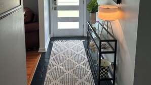 A hallway with a white door, a hanging light fixture, a potted plant, a console table with a lamp, and a patterned rug.