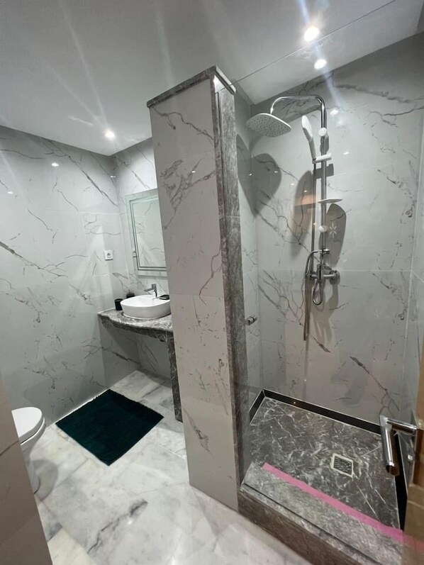 Un baño moderno con piso de mármol, cabina de ducha con paredes de vidrio y lavamanos sobre encimera de mármol.