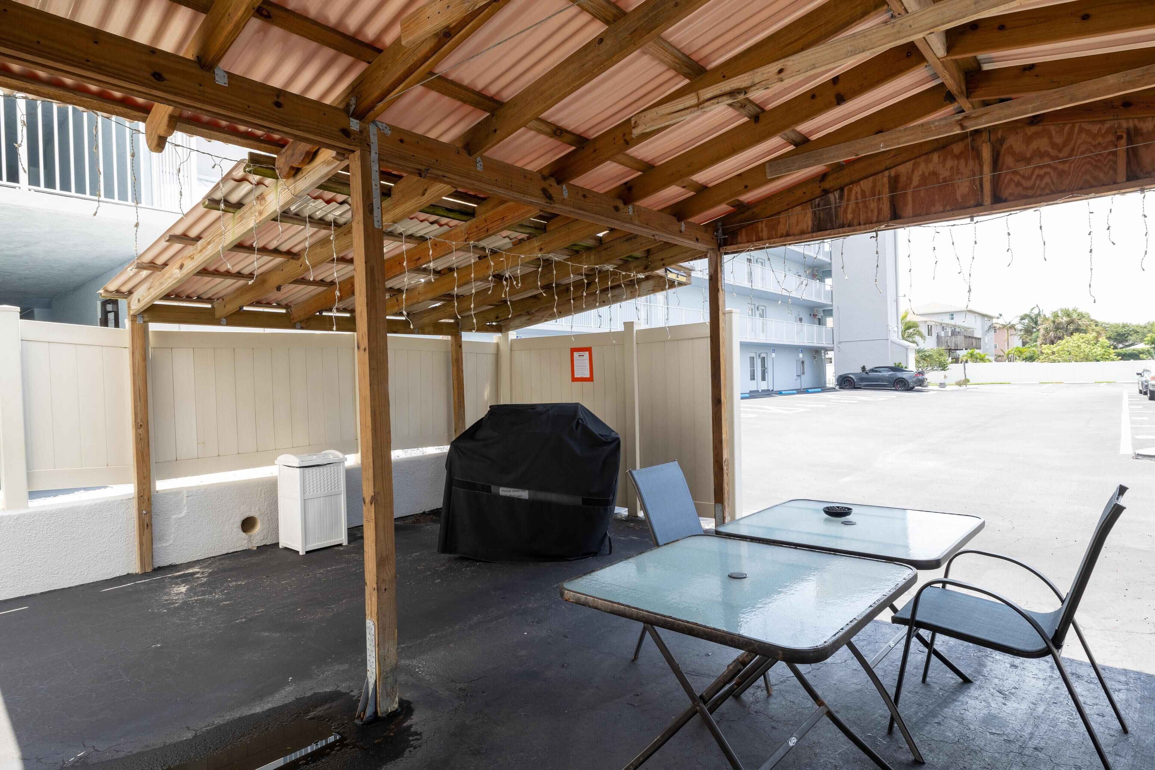 Een overdekt terras buiten met een tafel, stoelen, een barbecue en een witte vuilnisbak.