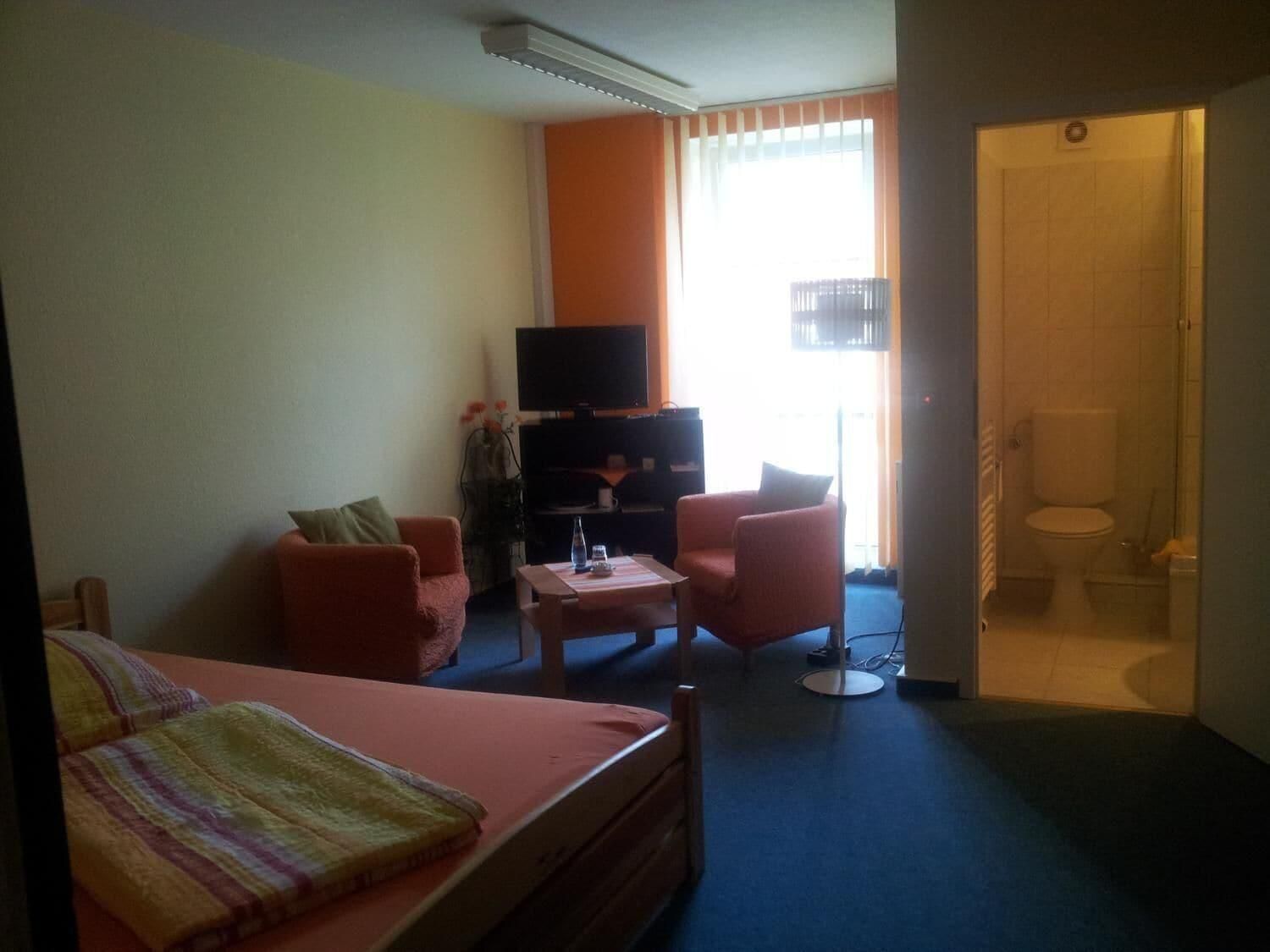 A hotel room with a bed, two armchairs, a small table, a TV, and a bathroom visible through an open door.