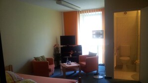 Habitación de hotel con cama, dos sillones, una mesita, televisor y baño visible a través de una puerta abierta.