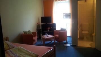 A hotel room with a bed, two armchairs, a small table, a TV, and a bathroom visible through an open door.