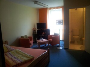 Habitación de hotel con cama, dos sillones, una mesita, televisor y baño visible a través de una puerta abierta.
