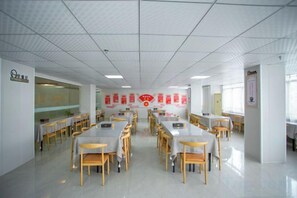 Un'ampia sala da pranzo con diversi tavoli e sedie, un soffitto piastrellato e segnaletica rossa alle pareti.