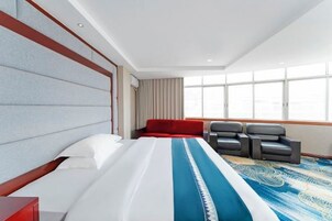 Habitación de hotel con una cama grande, un sillón rojo, un sofá gris y un ventanal con vistas.