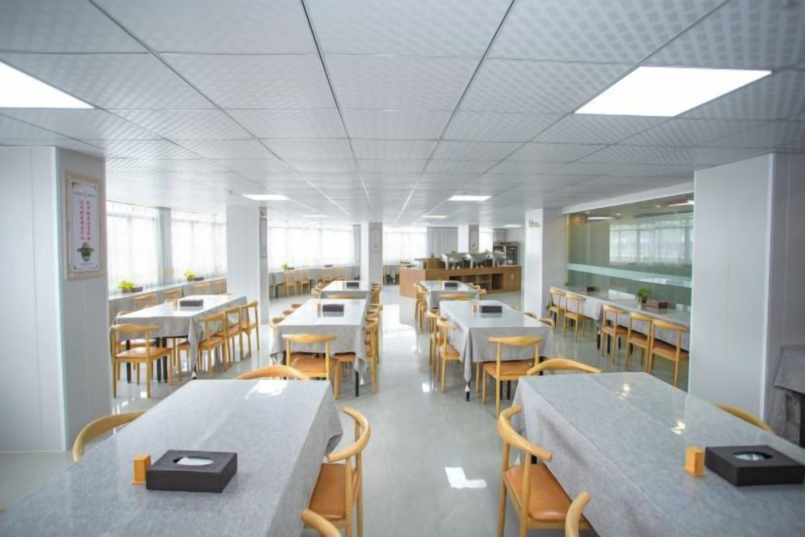 A spacious dining area with multiple tables and chairs, a kitchen in the background, and a tiled ceiling.