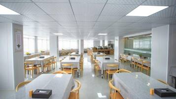 A spacious dining area with multiple tables and chairs, a kitchen in the background, and a tiled ceiling.