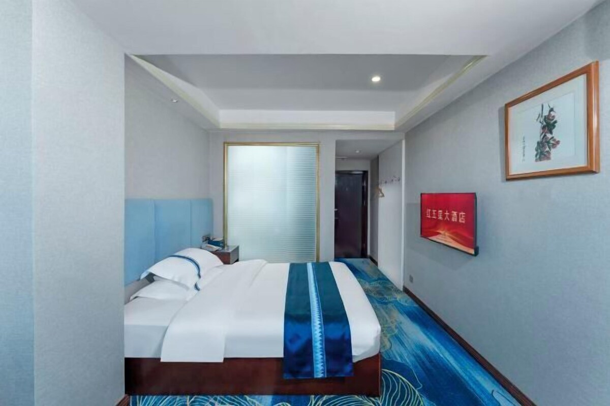 A hotel room with a bed, a nightstand, a window with blinds, a framed picture on the wall, and a carpet with a blue pattern.