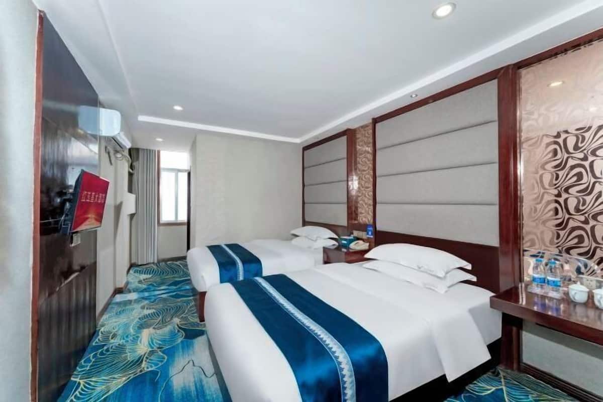 Une chambre d’hôtel avec deux lits, une tête de lit en bois, une moquette à motifs et une table de chevet avec des bouteilles d’eau.