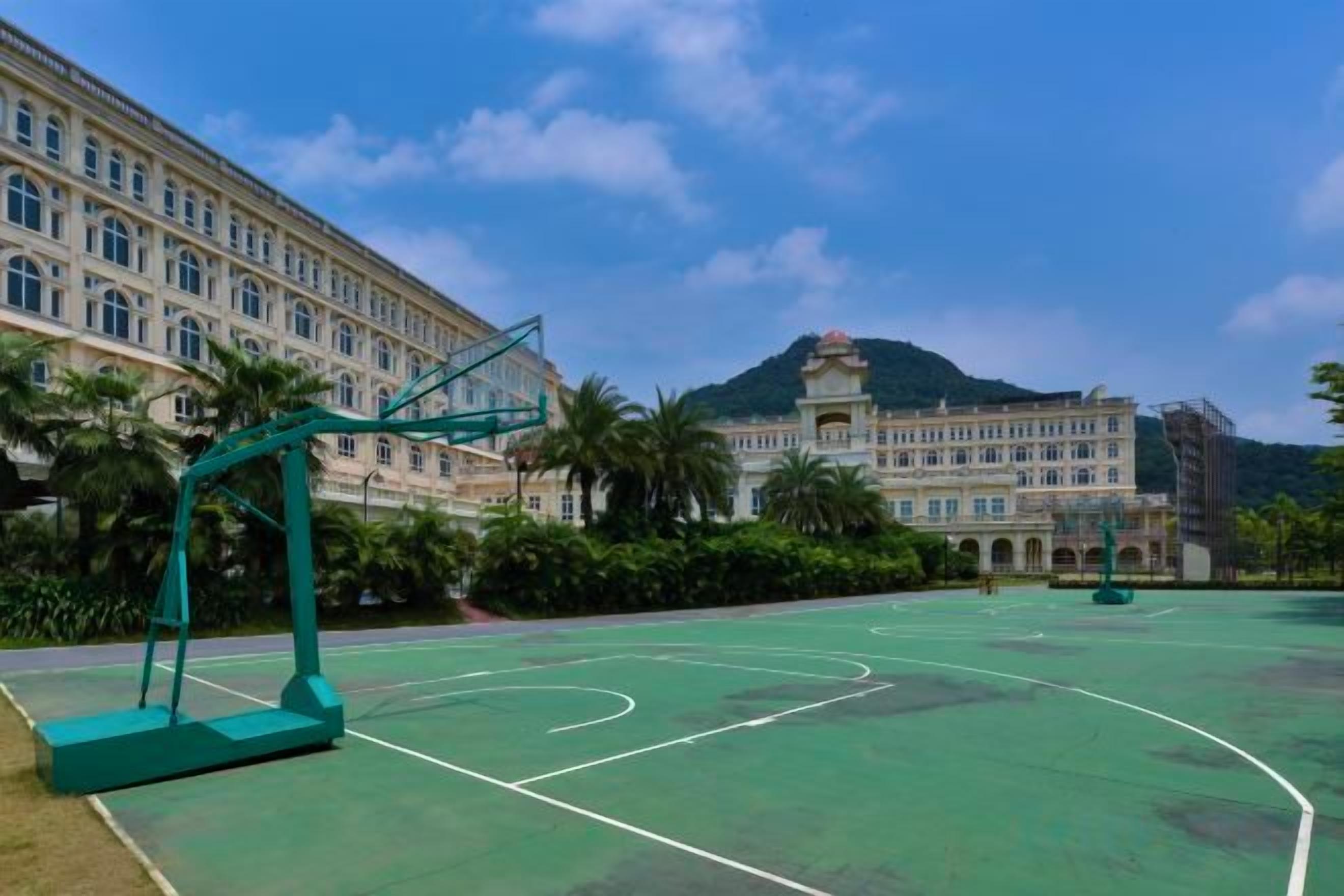 Un terrain de basket avec un filet vert, un bâtiment à l’architecture classique et une montagne en arrière-plan.