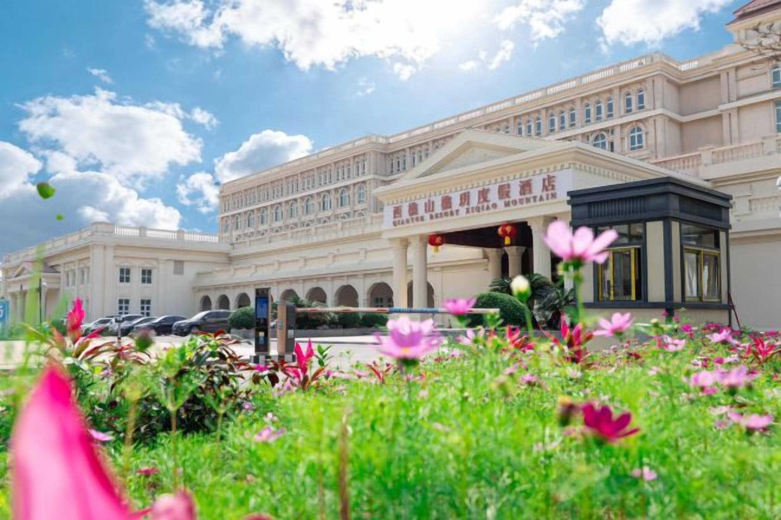 Ein großes Gebäude im klassischen Stil, mit einem zentralen Eingangsbereich und einer Portikus, umgeben von einem gepflegten Garten mit verschiedenen Pflanzen und Blumen.