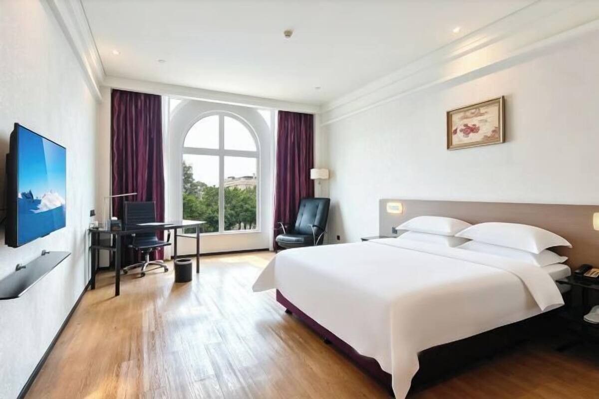 A hotel room with a large bed, a desk with a computer, a TV, and a window with curtains.