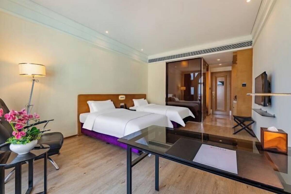 A hotel room with a bed, a desk, a chair, a TV, and a bathroom visible through a glass door.