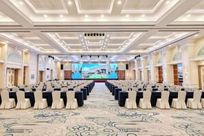 Une grande salle de conférence avec des rangées de tables et de chaises, un grand écran à l’avant et une moquette à motifs.