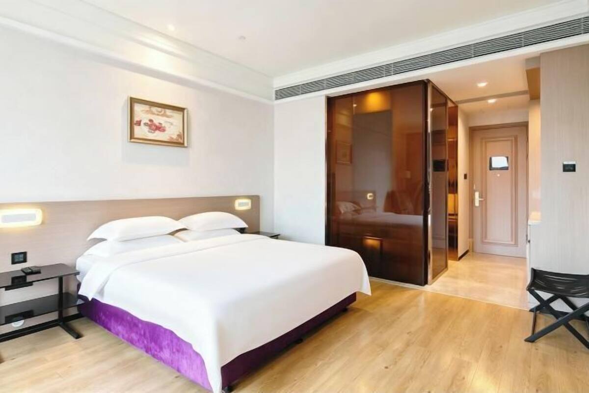 A modern hotel room with a large bed, a bedside table, a wardrobe, and a small desk.