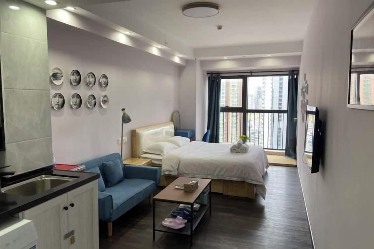 Een moderne hotelkamer met een bed, een blauwe bank, een kleine tafel, een bureau, een kitchenette en een groot raam met uitzicht op de stad.