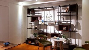 Une pièce moderne avec une table à manger en bois, une chaise verte, une petite table ronde recouverte d’une nappe blanche et une bibliothèque en métal noir remplie de livres et d’objets décoratifs.