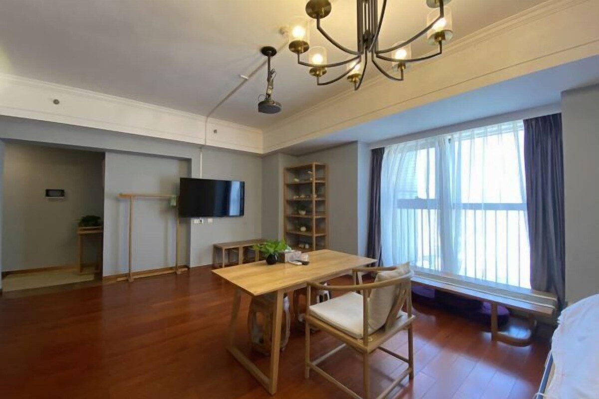 Ein geräumiges Zimmer mit Holzboden, einem Esstisch mit Stühlen, einem Fernseher, einem Bücherregal und einem großen Fenster mit Vorhängen.