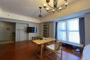 Ein geräumiges Zimmer mit Holzboden, einem Esstisch mit Stühlen, einem Fernseher, einem Bücherregal und einem großen Fenster mit Vorhängen.