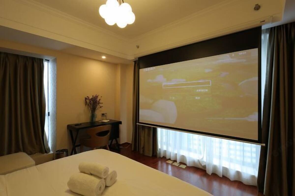 Hotelkamer met een bed, bureau, stoel, projectiescherm en raam met gordijnen.