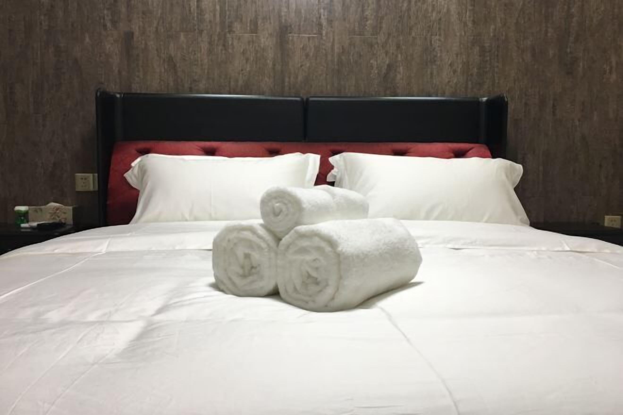 A neatly made bed with white linens and rolled towels, a dark headboard, and a wooden wall.
