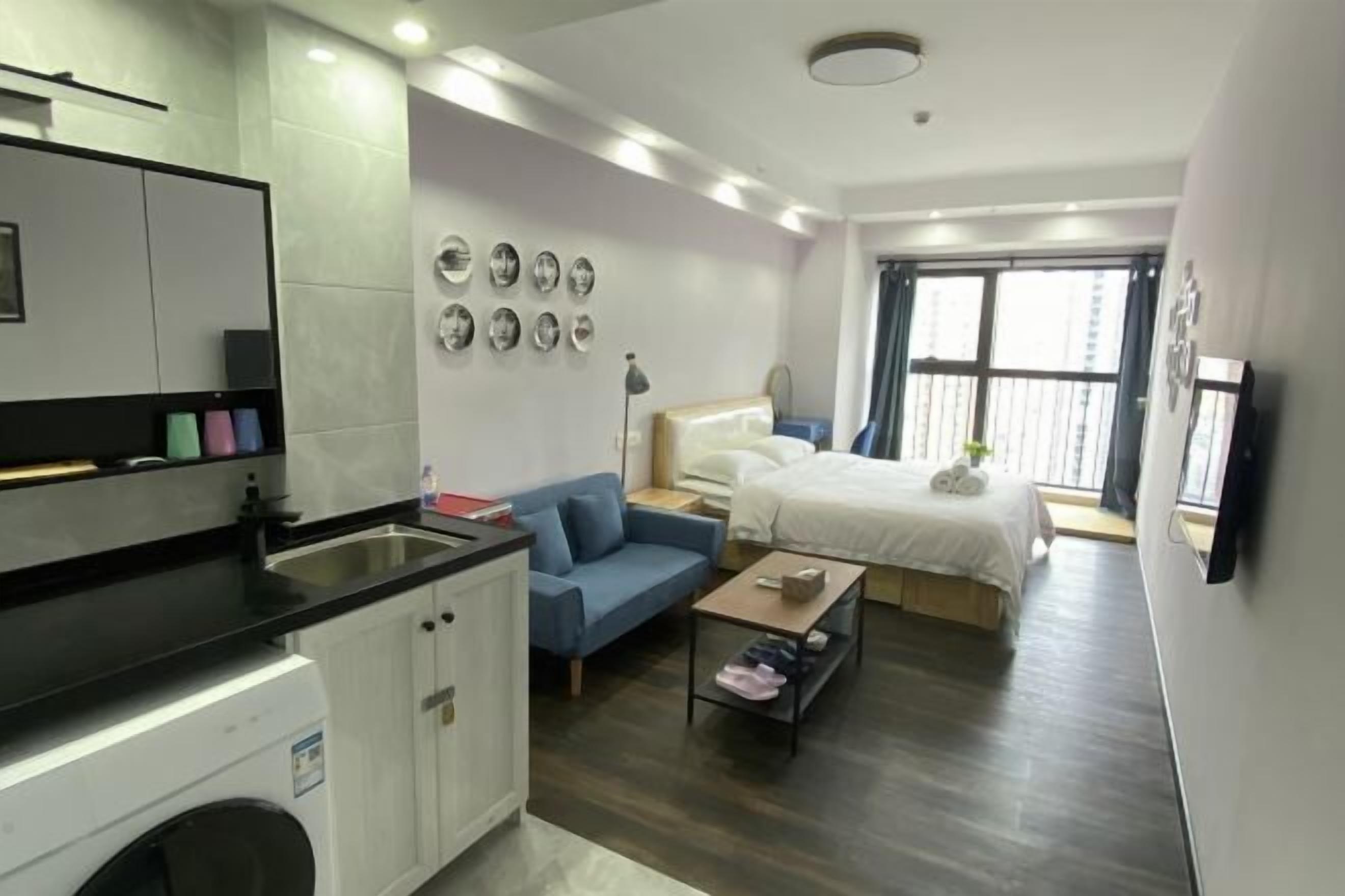 A compact hotel room with a kitchenette, a bed, a sofa, a small table, and a window with a view.