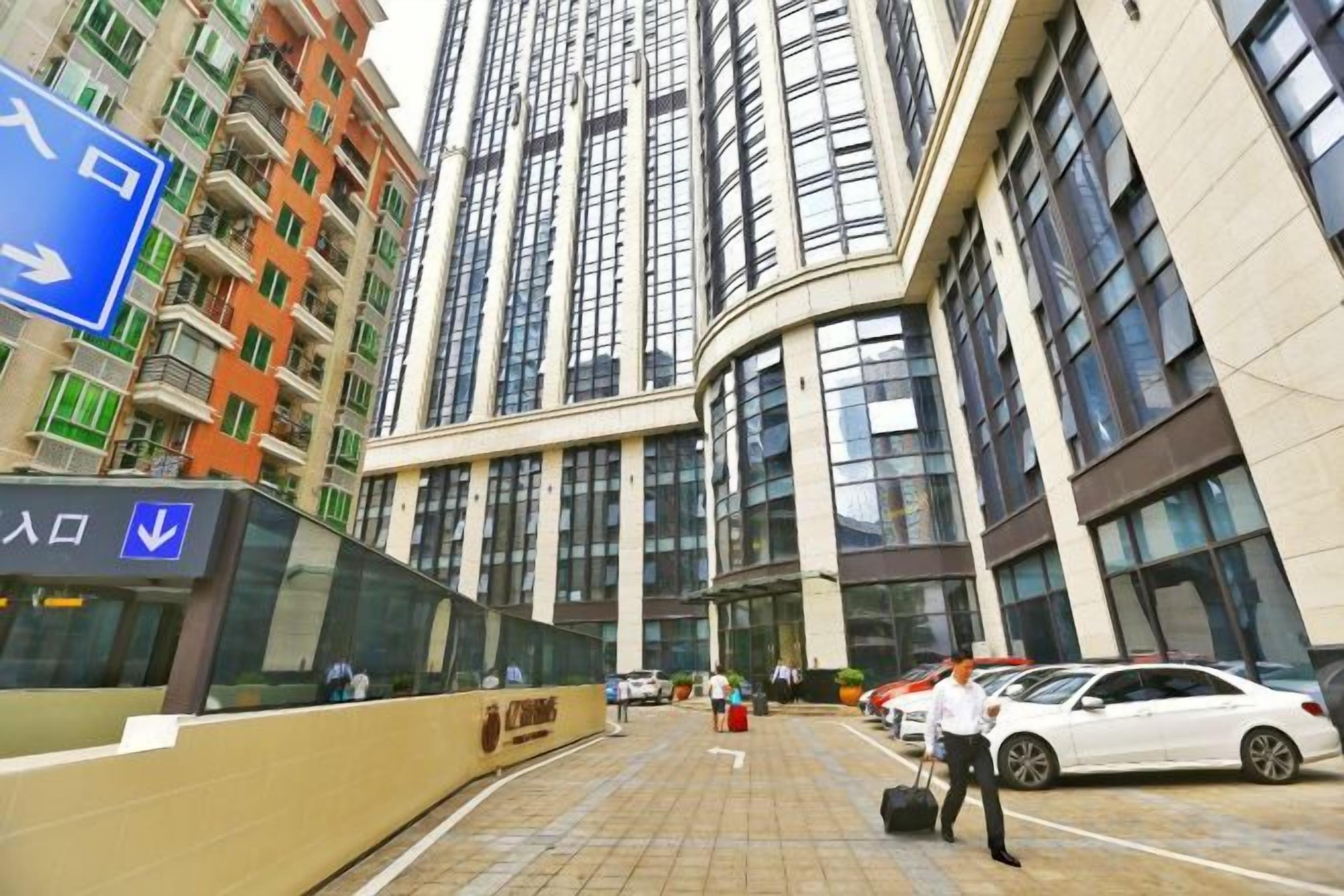 Un edificio de gran altura con diseño moderno, que cuenta con una entrada destacada y un estacionamiento con coches.