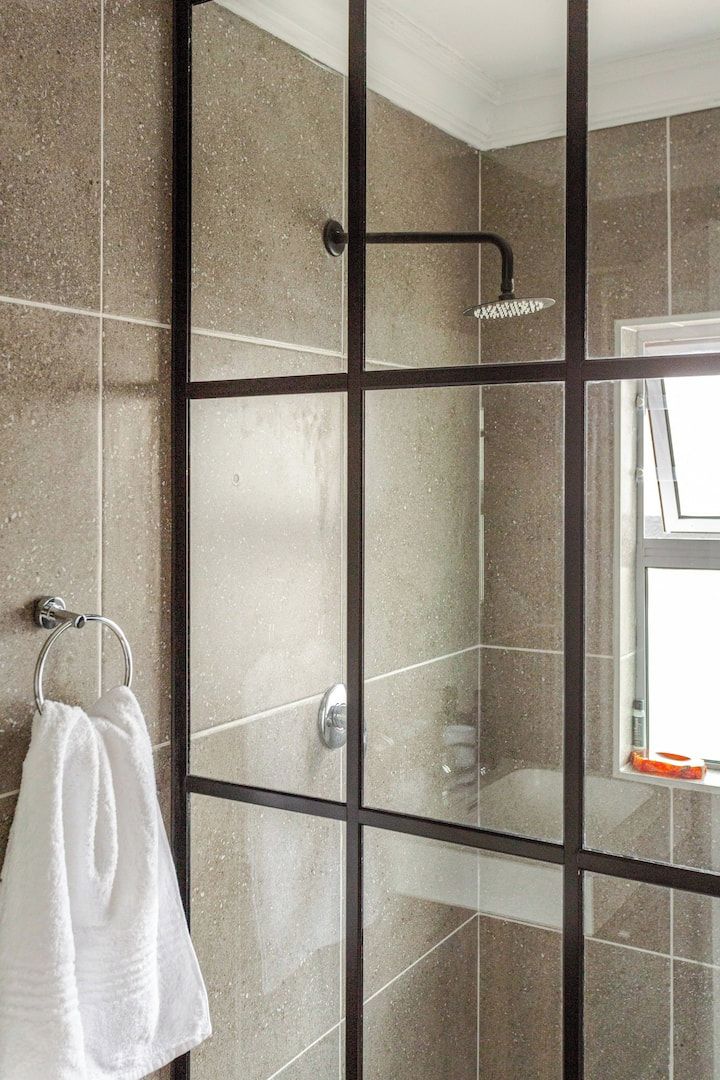 A glass-enclosed shower with a handheld showerhead, a towel rack with a white towel, and a window reflecting a bright interior.