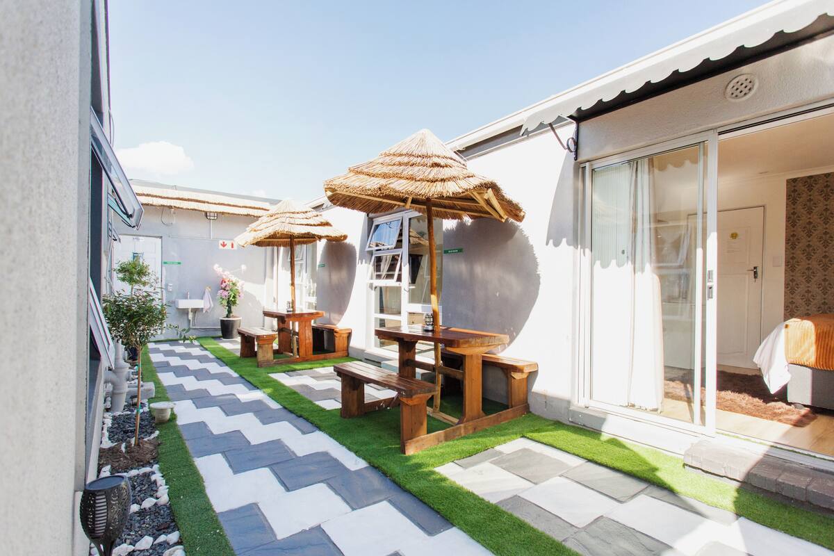 A patio with wooden tables and thatched umbrellas, a small garden, and a building with a sliding glass door.