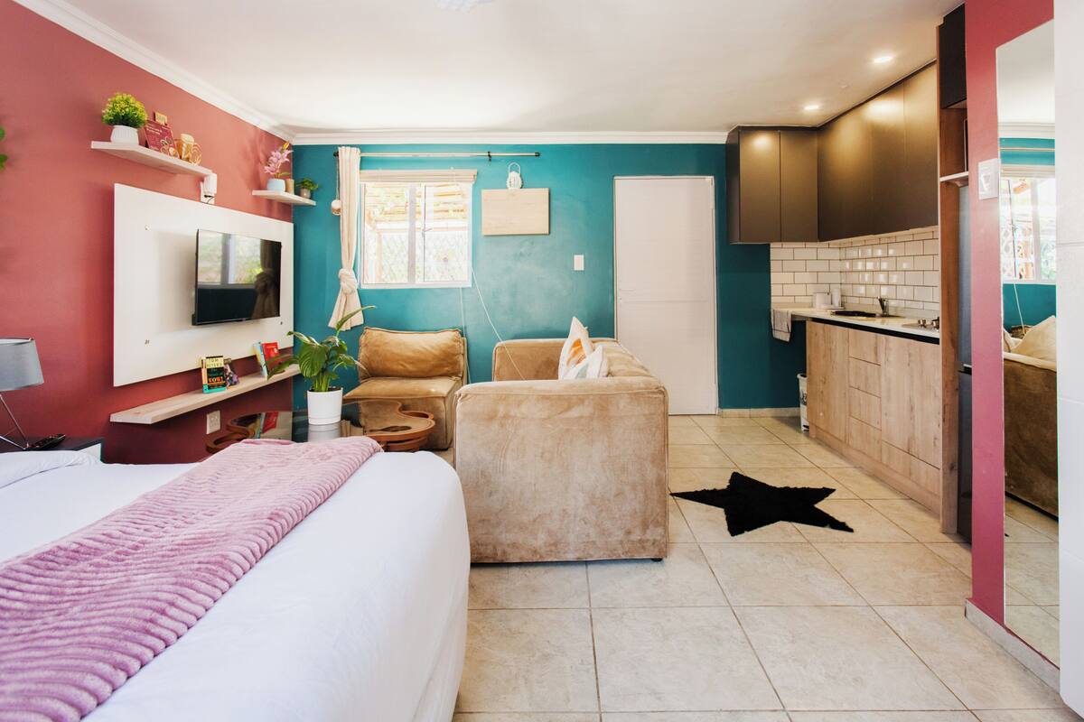 A modern hotel room with a bed, a sofa, a TV, a kitchen area, and a bathroom.